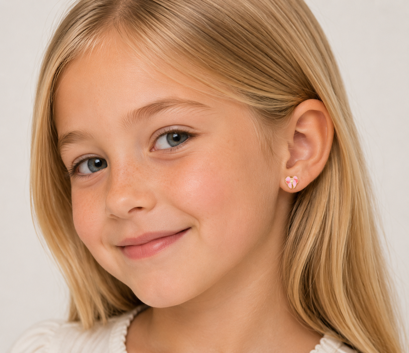 A young girl with long blonde hair smiles softly, wearing JaxKelly's Bow Enamel Flat Back Kids Earrings—hypoallergenic and perfect for sensitive ears. The neutral background highlights her face.
