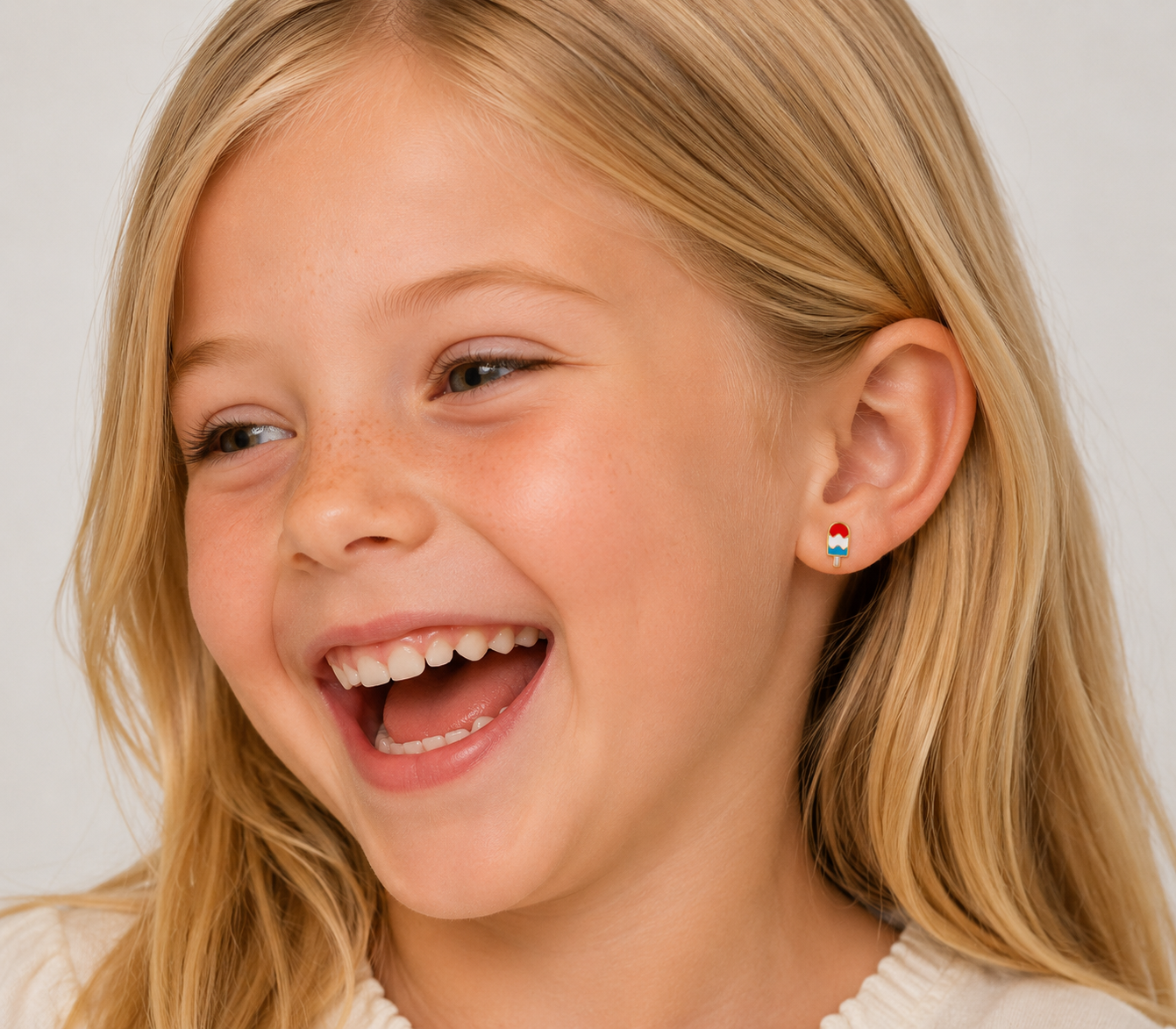 A young blonde girl with long hair smiles joyfully, showing her teeth. She wears a cream top and JaxKelly Popsicle Enamel Flat Back Kids Earrings on her left ear. A plain light background highlights her playful accessory.