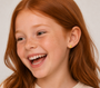 A young girl with long red hair and freckles smiles joyfully, wearing a light-colored top and JaxKelly Pizza Enamel Flat Back Kids Earrings. Her small, yellow pizza-shaped earrings stand out against the plain, neutral background.