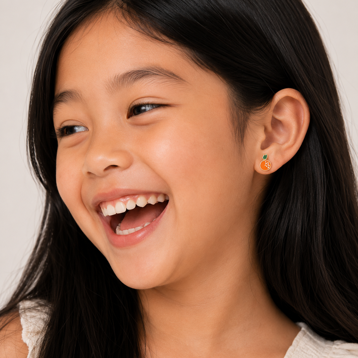 A young girl with long black hair smiles joyfully, wearing a white top and JaxKelly Orange Enamel Flat Back Kids Earrings made from surgical-grade stainless steel. The softly lit background is neutral.