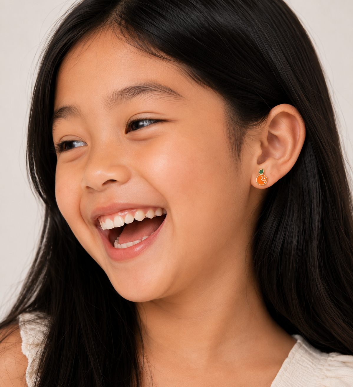 A young girl with long black hair smiles joyfully, wearing a white top and JaxKelly Orange Enamel Flat Back Kids Earrings made from surgical-grade stainless steel. The softly lit background is neutral.