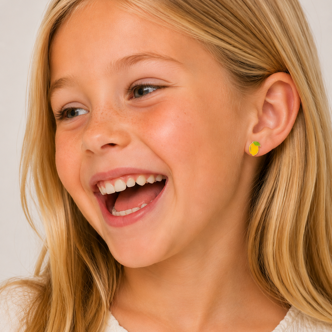 A young girl with long blonde hair smiles joyfully, wearing a cream top and JaxKelly Lemon Enamel Flat Back Kids Earrings. The background is light and neutral.