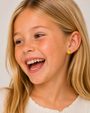 A young girl with long blonde hair smiles joyfully, wearing a cream top and JaxKelly Lemon Enamel Flat Back Kids Earrings. The background is light and neutral.