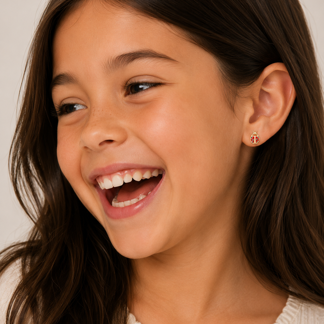 A young girl with long brown hair smiles brightly, wearing a cream-colored top and JaxKelly Ladybug Enamel Flat Back Kids Earrings made from hypoallergenic, surgical-grade stainless steel. The softly lit background is neutral.