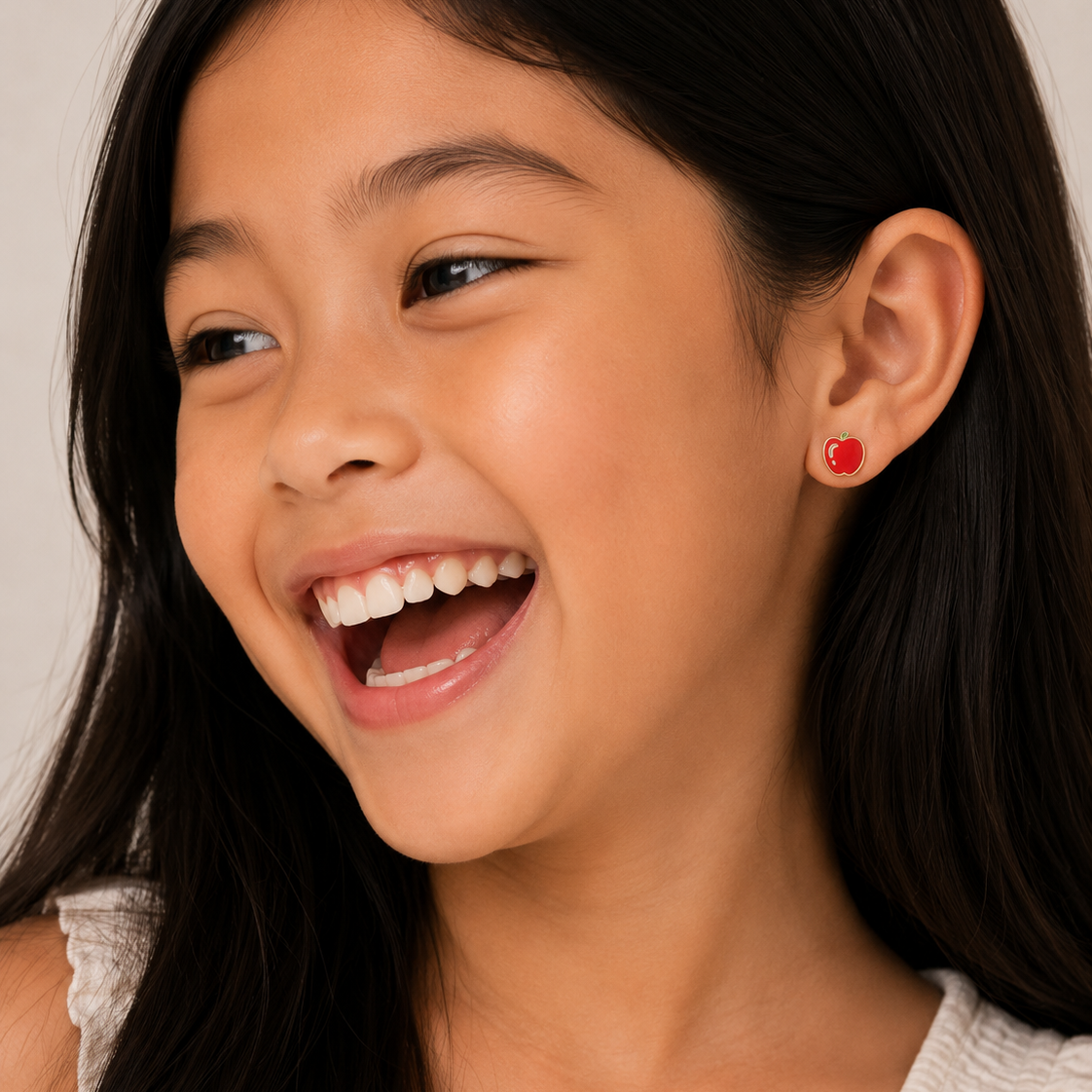 A young girl with long dark hair smiles brightly, wearing a light top and JaxKelly’s Apple Enamel Flat Back Kids Earrings made from hypoallergenic, surgical-grade stainless steel. The softly blurred background is neutral.
