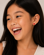 A young girl with long dark hair smiles brightly, wearing a light top and JaxKelly’s Apple Enamel Flat Back Kids Earrings made from hypoallergenic, surgical-grade stainless steel. The softly blurred background is neutral.