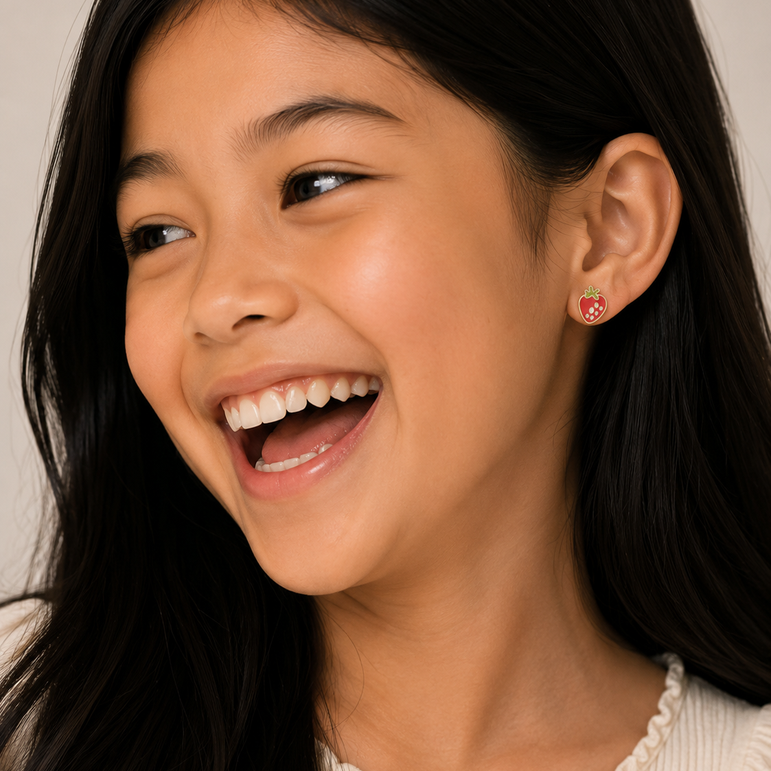 A young girl with long dark hair smiles, looking to the side. She wears a cream top and JaxKelly Strawberry Enamel Flat Back Kids Earrings—small, round, hypoallergenic studs perfect for sensitive ears.