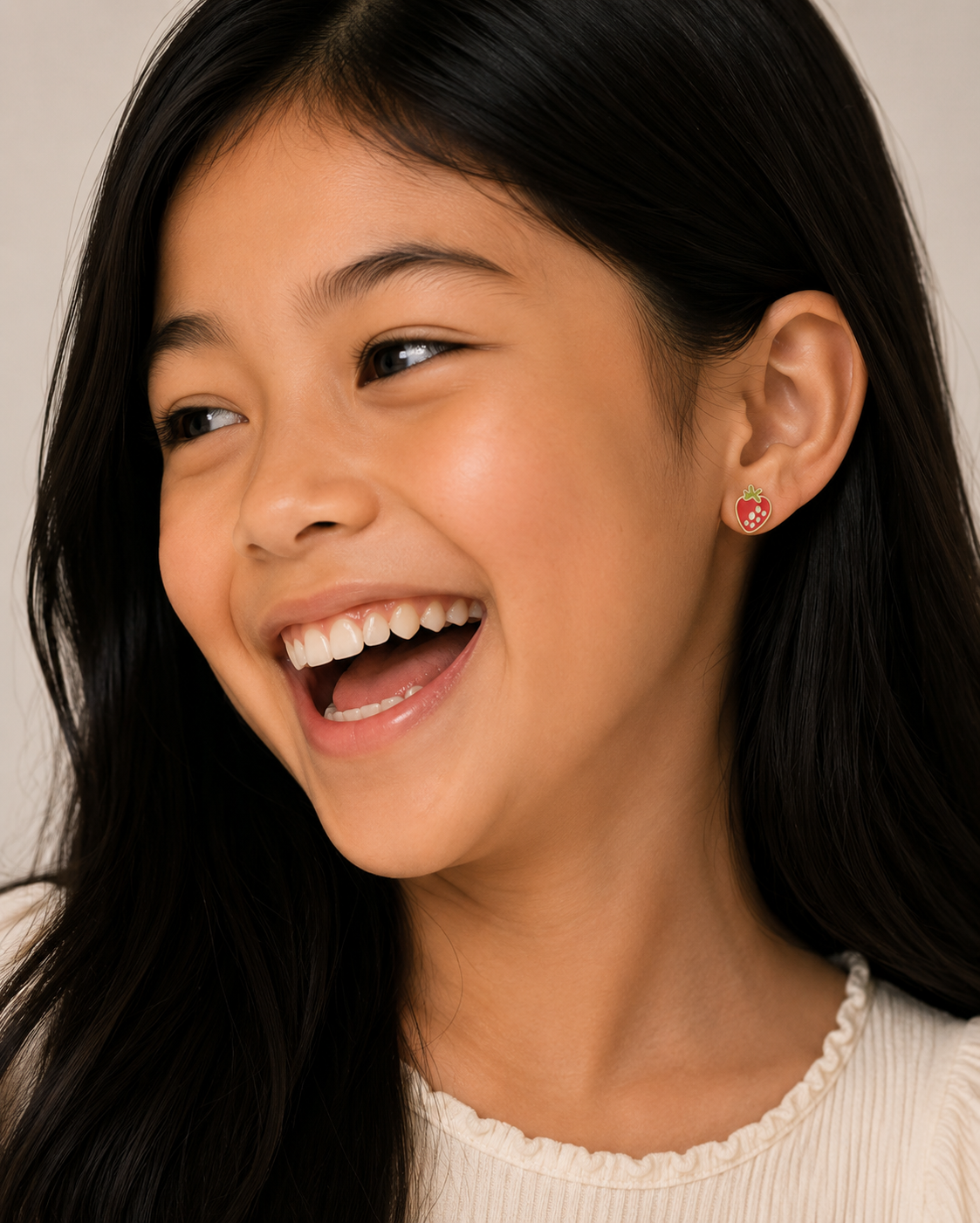 A young girl with long dark hair smiles, looking to the side. She wears a cream top and JaxKelly Strawberry Enamel Flat Back Kids Earrings—small, round, hypoallergenic studs perfect for sensitive ears.