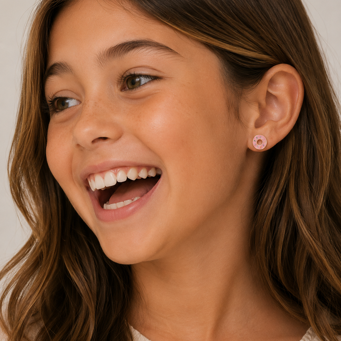 A young girl with long brown hair smiles brightly, glancing to the side. She wears a light top and JaxKelly Donut Enamel Flat Back Kids Earrings. The neutral background is softly lit.