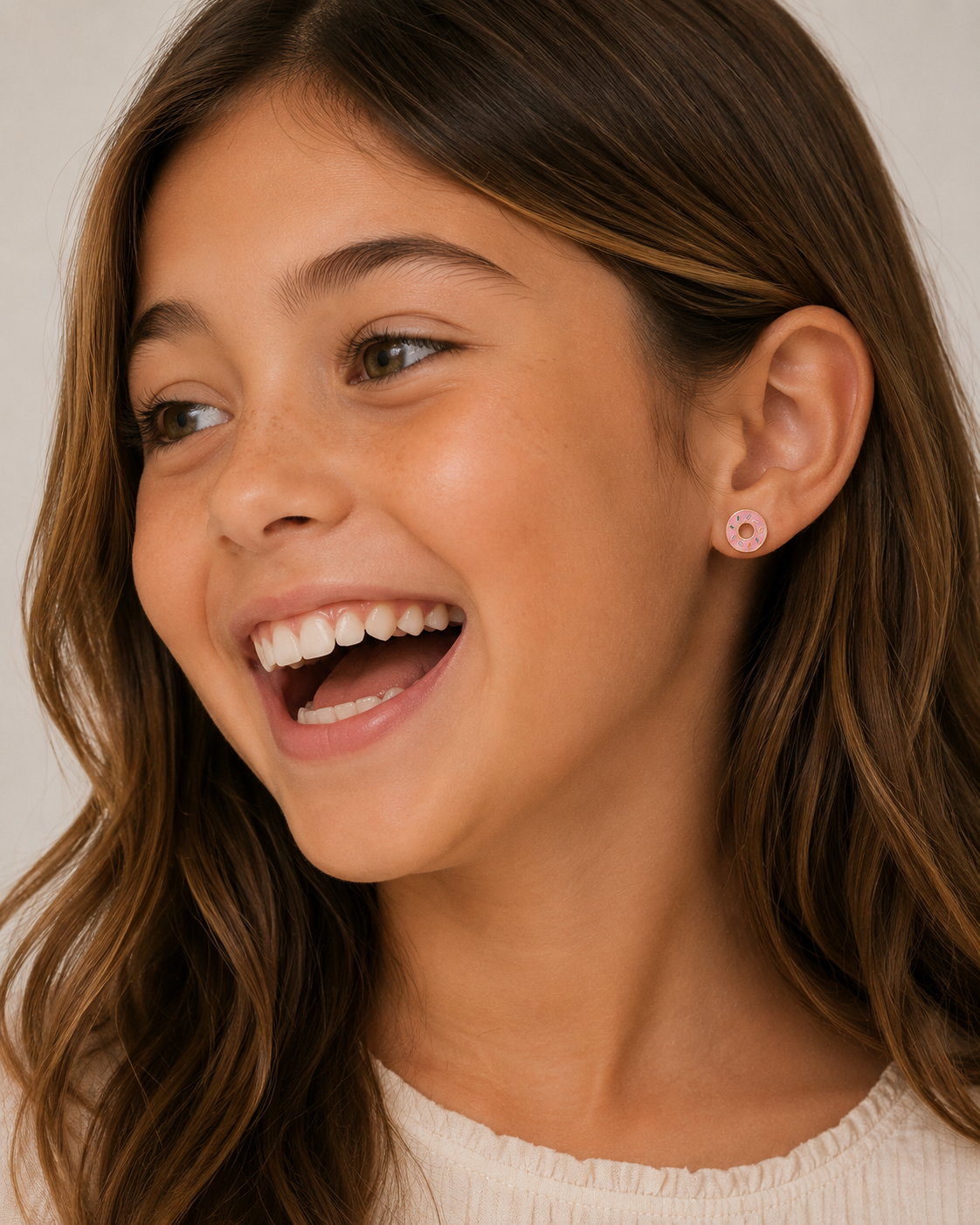 A young girl with long brown hair smiles brightly, glancing to the side. She wears a light top and JaxKelly Donut Enamel Flat Back Kids Earrings. The neutral background is softly lit.