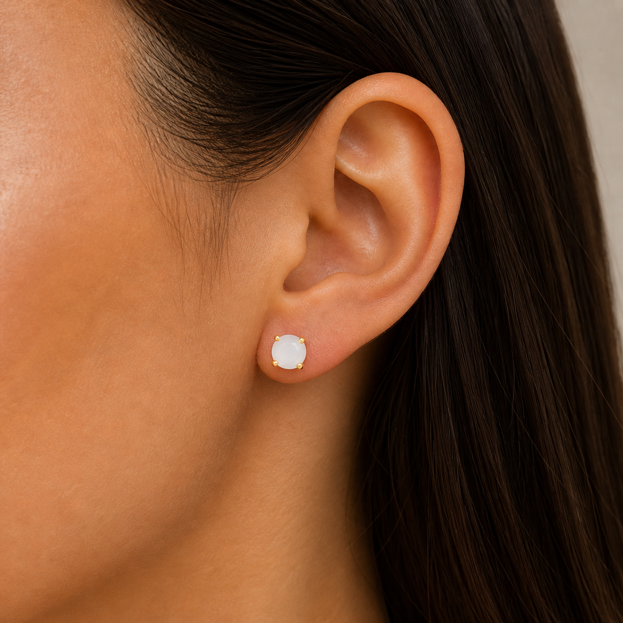 A close-up shows a woman with smooth, medium-toned skin and straight brown hair wearing JaxKelly’s Round Mother of Pearl Gemstone Gold Earrings.