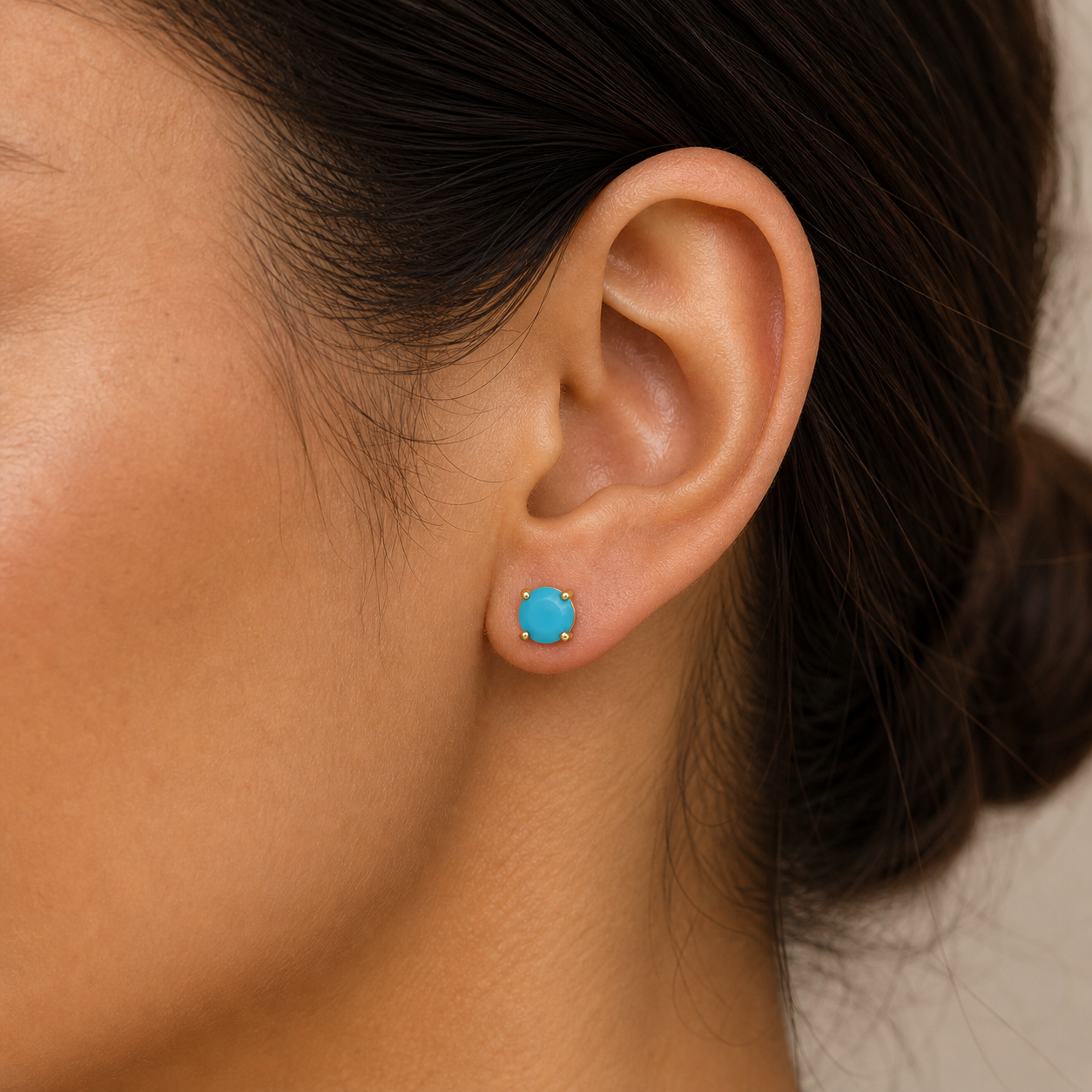 A woman wears JaxKelly's Round Turquoise Gemstone Gold Earrings; her hair is tied back to showcase the minimalist turquoise studs and gold-plated setting, highlighting their elegant design against her natural complexion.
