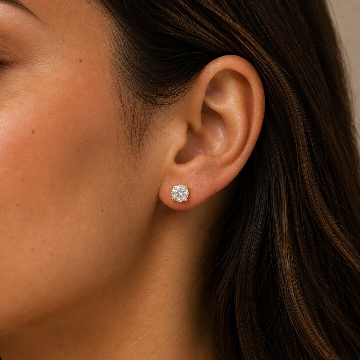 Close-up of a woman's ear adorned with JaxKelly Round CZ - Clear - Gold Earrings. Her brown hair is tucked behind her ear, highlighting the minimalist gold-plated studs against her warm, natural skin tone.