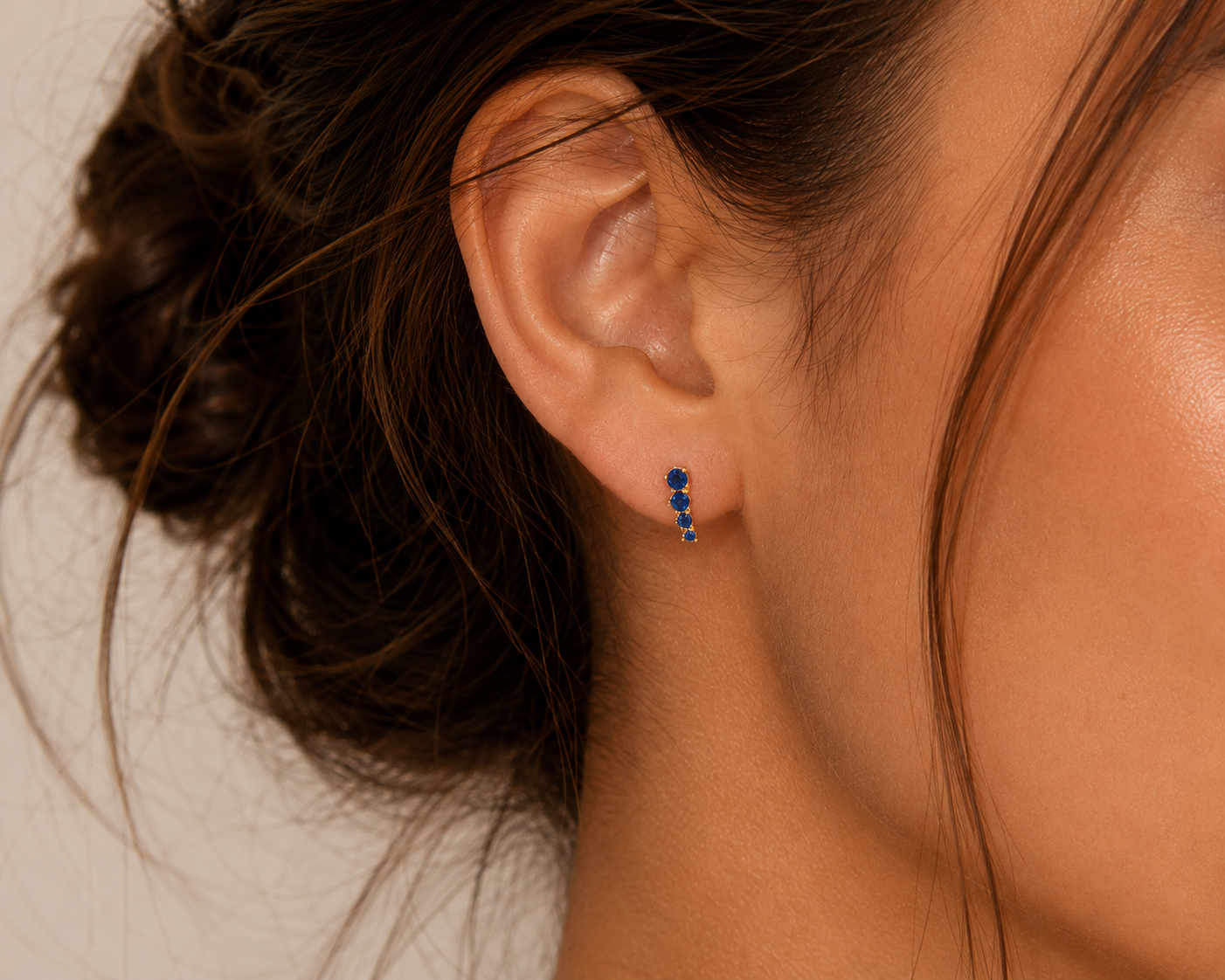 A close-up shows a woman’s ear wearing JaxKelly’s Mini Crawler - Sapphire CZ - Gold Earrings; her loose bun highlights the elegant vertical row design against her warm, natural-toned skin.