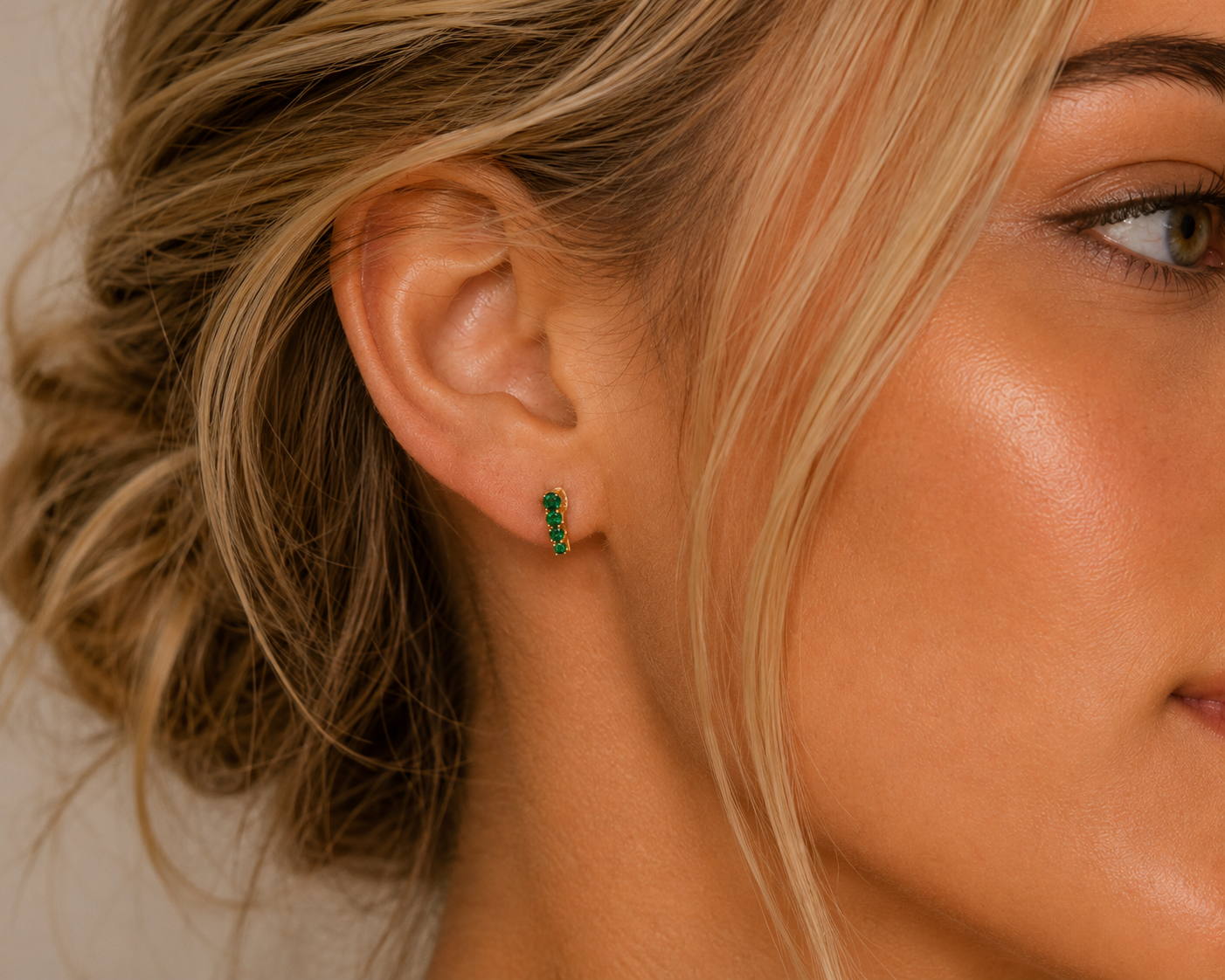 A close-up shows a woman's ear wearing JaxKelly's Mini Crawler - Emerald CZ - Gold Earrings. Her blonde hair is in an elegant updo, with tendrils framing her face and lighting accentuating her glowing skin.