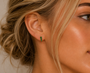 A close-up shows a woman's ear wearing JaxKelly's Mini Crawler - Emerald CZ - Gold Earrings. Her blonde hair is in an elegant updo, with tendrils framing her face and lighting accentuating her glowing skin.