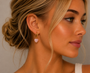 A woman with blonde hair in a loose updo wears JaxKelly's Heart Drop - Rose Quartz tarnish-free gold hoop earrings and a delicate gold necklace, pairing them with her white top and natural makeup as she looks to the right.