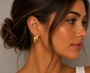 A woman with tan skin and brown hair in a loose bun wears the Gold Double Bold Bar earrings by JaxKelly, shown in profile against a neutral background. She features natural makeup, and the earrings have a soft, tarnish-free shine.