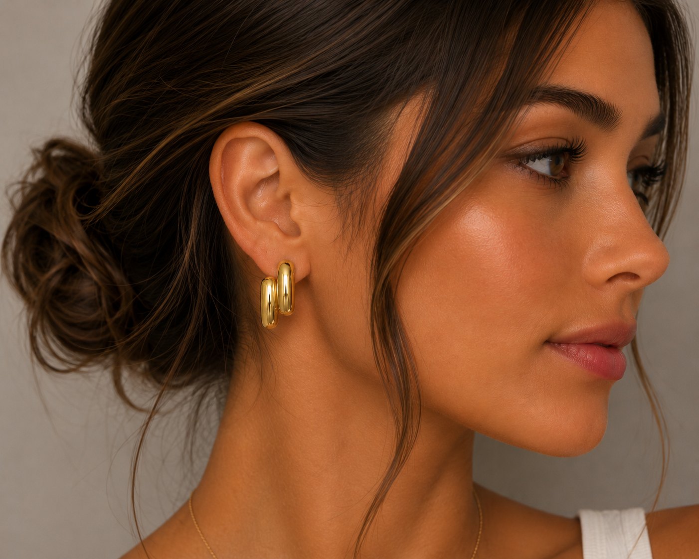 A woman with tan skin and brown hair in a loose bun wears the Gold Double Bold Bar earrings by JaxKelly, shown in profile against a neutral background. She features natural makeup, and the earrings have a soft, tarnish-free shine.