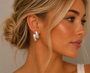 A woman with blonde hair in a loose bun wears JaxKelly's Silver Double Bold Bar tarnish-free silver hoop earrings with sterling silver posts and a gold necklace, gazing to the side. Her skin glows as she dons a light-colored top.