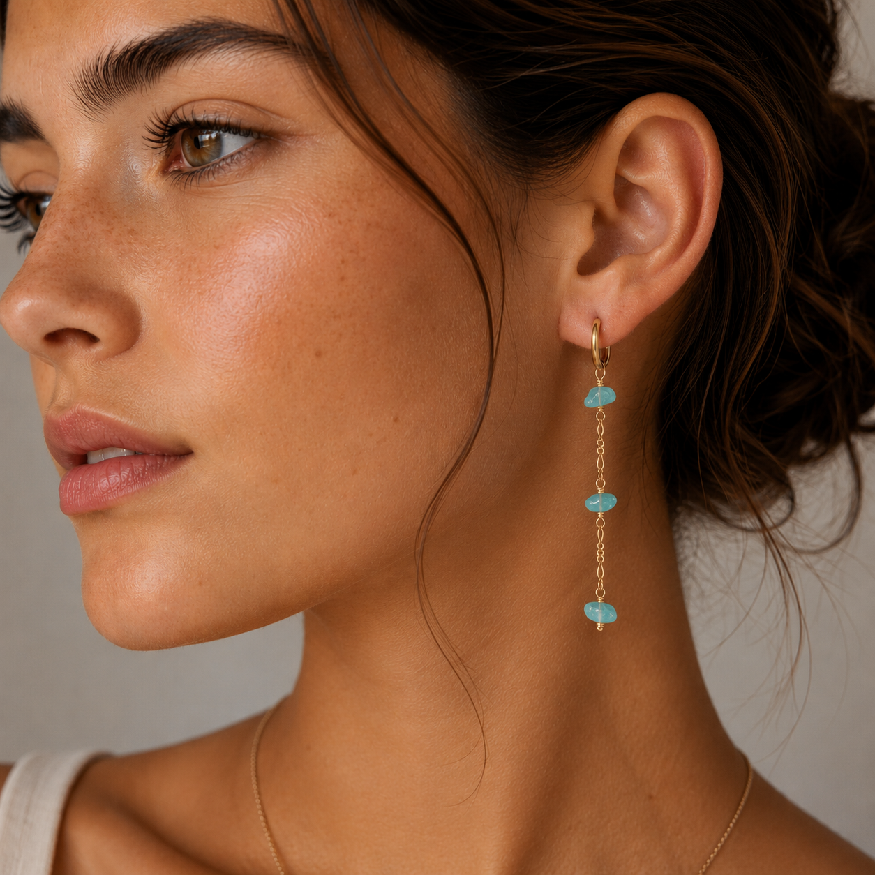 A woman with glowing skin and brown hair wears JaxKelly Raw Gemstone Chain Drop Earrings in amazonite, featuring turquoise stones and a simple gold necklace. Shown in profile against a neutral background, her elegant earrings stand out.