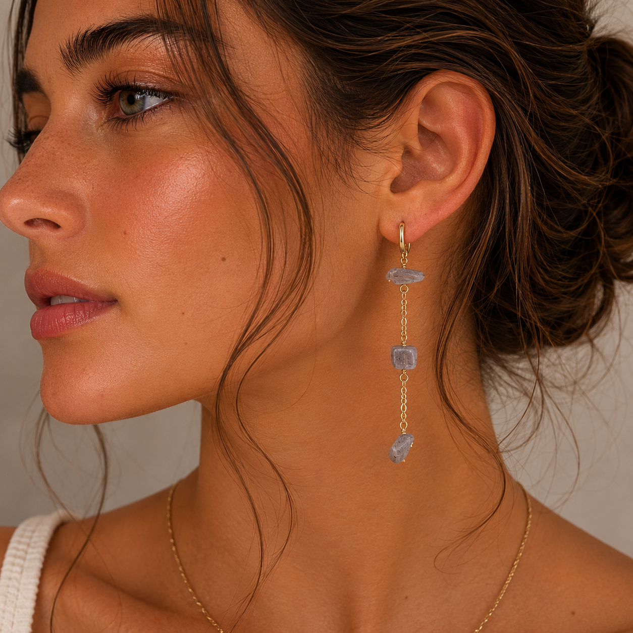 A woman with wavy brown hair wears JaxKelly's Raw Gemstone Chain Drop Earrings in rainbow moonstone and a delicate gold necklace. She looks to the side, her skin glowing with natural makeup.