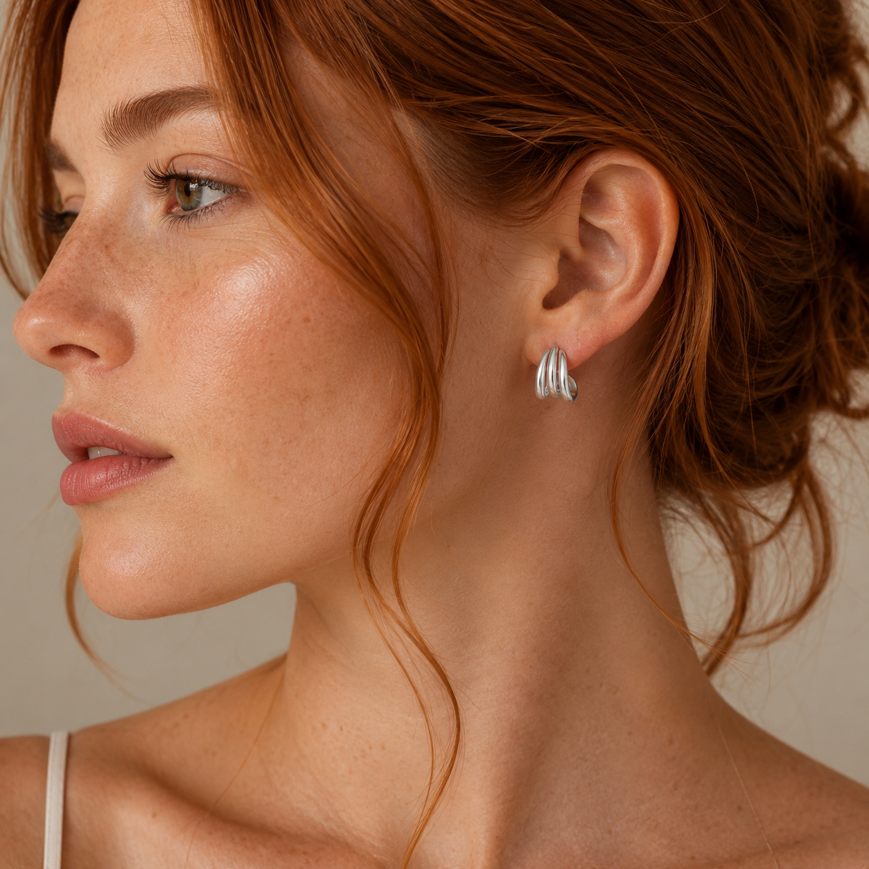 A woman with fair skin and loose red waves is seen in profile, wearing the JaxKelly Silver Three Hoop Huggie earrings with sterling silver posts, minimal makeup, a white spaghetti strap top, and a calm expression—minimalist jewelry perfected.