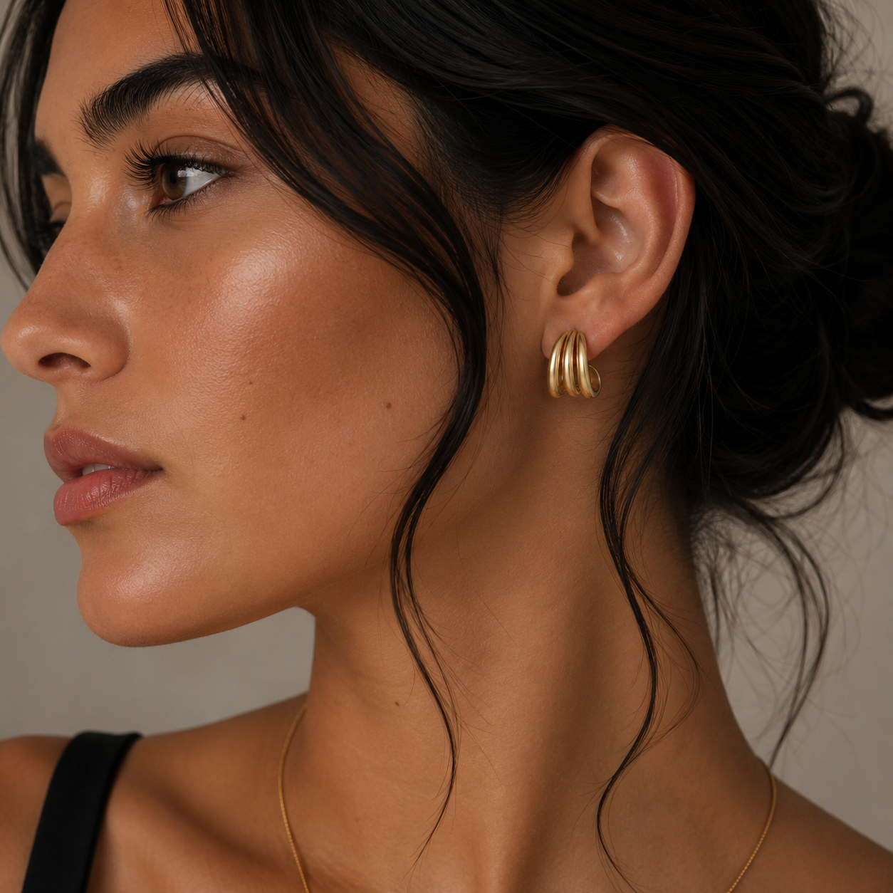 A woman with smooth, glowing skin and dark hair in a loose updo wears the JaxKelly Gold Three Hoop Huggie earrings and a thin gold necklace, looking off to the side as soft lighting enhances her natural features.