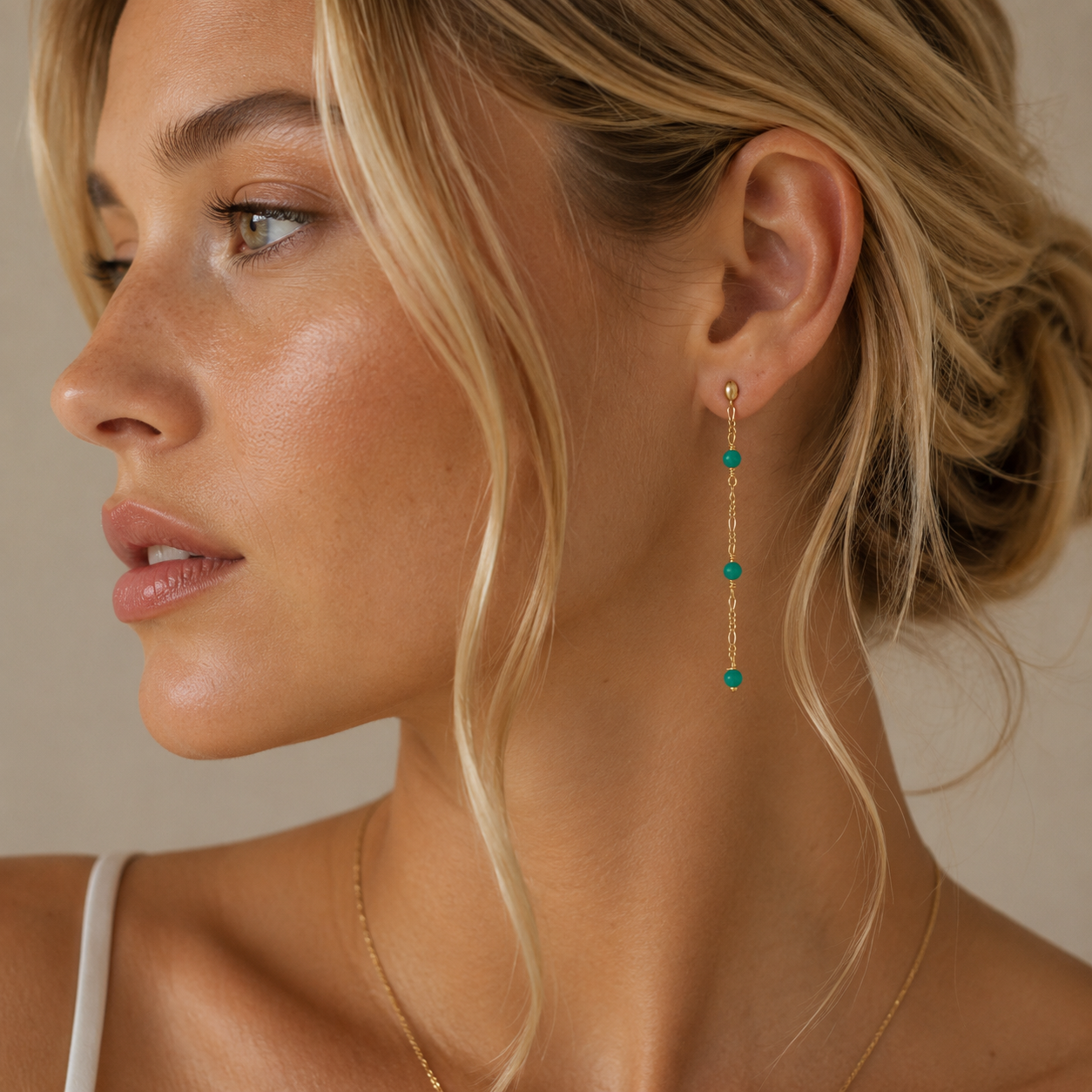 A woman with blonde hair in a loose updo wears JaxKelly's Gemstone Chain Drop - Turquoise earrings with turquoise beads and a delicate necklace. Shown in profile, she looks left, wearing a white top against a neutral background.