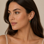 A woman with long brown hair looks to the side, wearing a beige top and JaxKelly's Gemstone Mini Drop - Malachite: tarnish-free gold plated hoops with green rectangular stones, against a plain light-colored background.