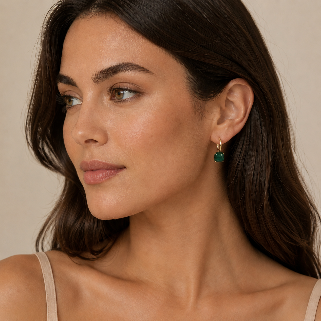A woman with long brown hair looks to the side, wearing a beige top and JaxKelly's Gemstone Mini Drop - Malachite: tarnish-free gold plated hoops with green rectangular stones, against a plain light-colored background.