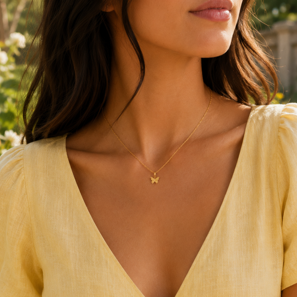 A woman outdoors in sunlight wears a pale yellow V-neck dress and the JaxKelly Mini Butterfly Pendant Necklace, an 18k gold plated piece with a diamond-cut chain.