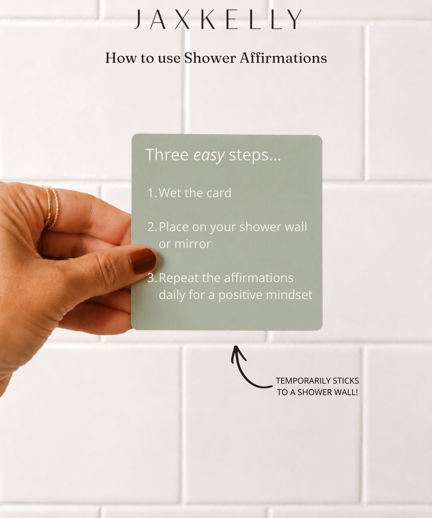 A hand with painted nails holds a green Strength Shower Affirmations™ card by JaxKelly, featuring waterproof instructions for use against a white tiled wall. Above, text reads "How to use Shower Affirmations" with an arrow pointing to the wall.