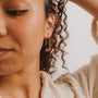 Close-up of a woman with curly hair in a cream textured top wearing the Mystery Earring—a gold hoop with pink crystal pendant—set against a neutral background. The Mystery Earring is part of our final sale collection.