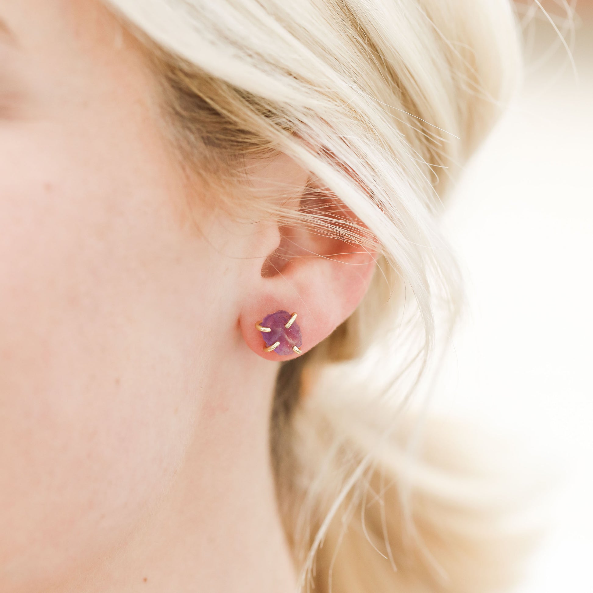 Close-up of a woman's ear wearing JaxKelly's Amethyst Gemstone Prong - Healing earring, a light purple stud set in gold plating. Her blonde hair and fair skin highlight the delicate design against an elegantly blurred background.