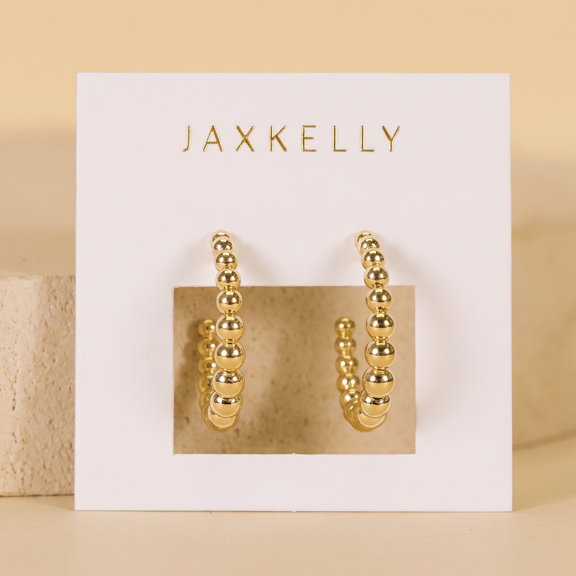 A pair of hypoallergenic Large Ball Hoop earrings by JaxKelly is displayed on a white card with the brand’s gold lettering, set against a neutral beige background.