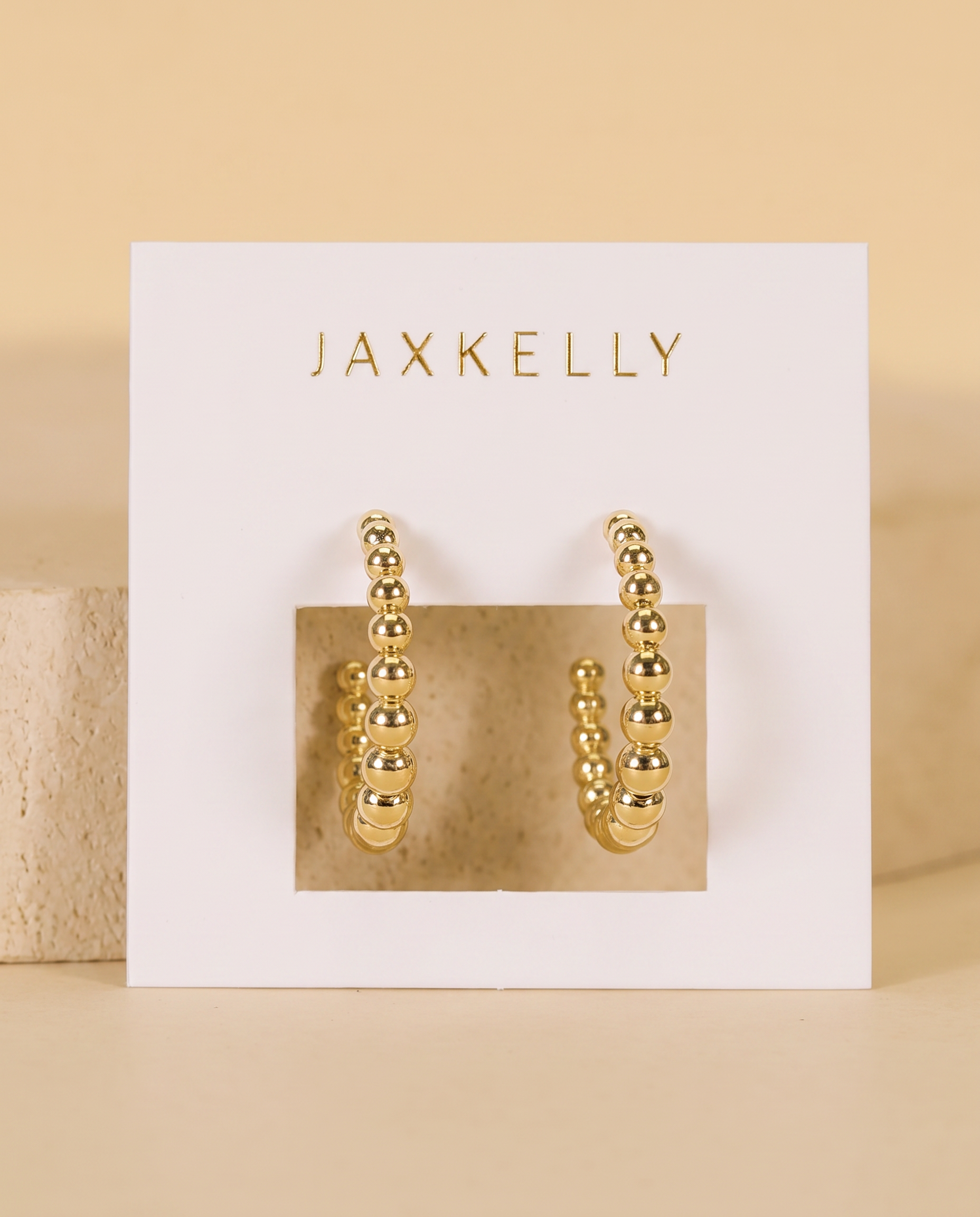 A pair of hypoallergenic Large Ball Hoop earrings by JaxKelly is displayed on a white card with the brand’s gold lettering, set against a neutral beige background.