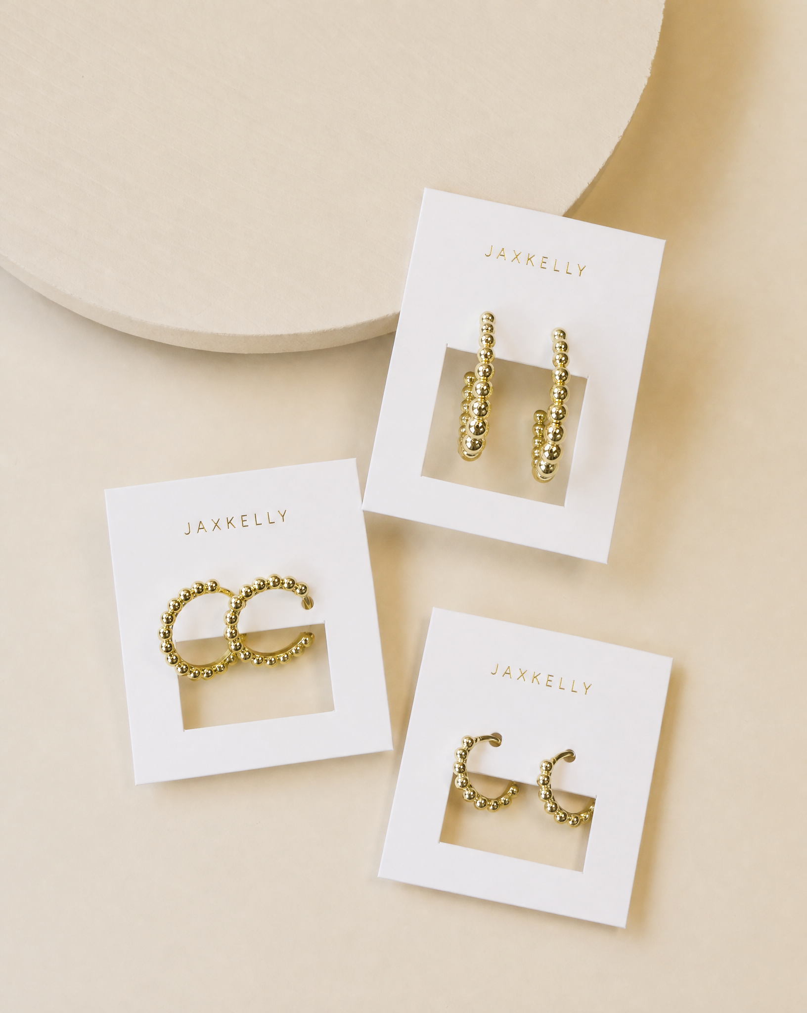Three pairs of JaxKelly Small Ball Hoop earrings are displayed on white branded cards, set on a beige surface with a round textured object partially visible at the top left.