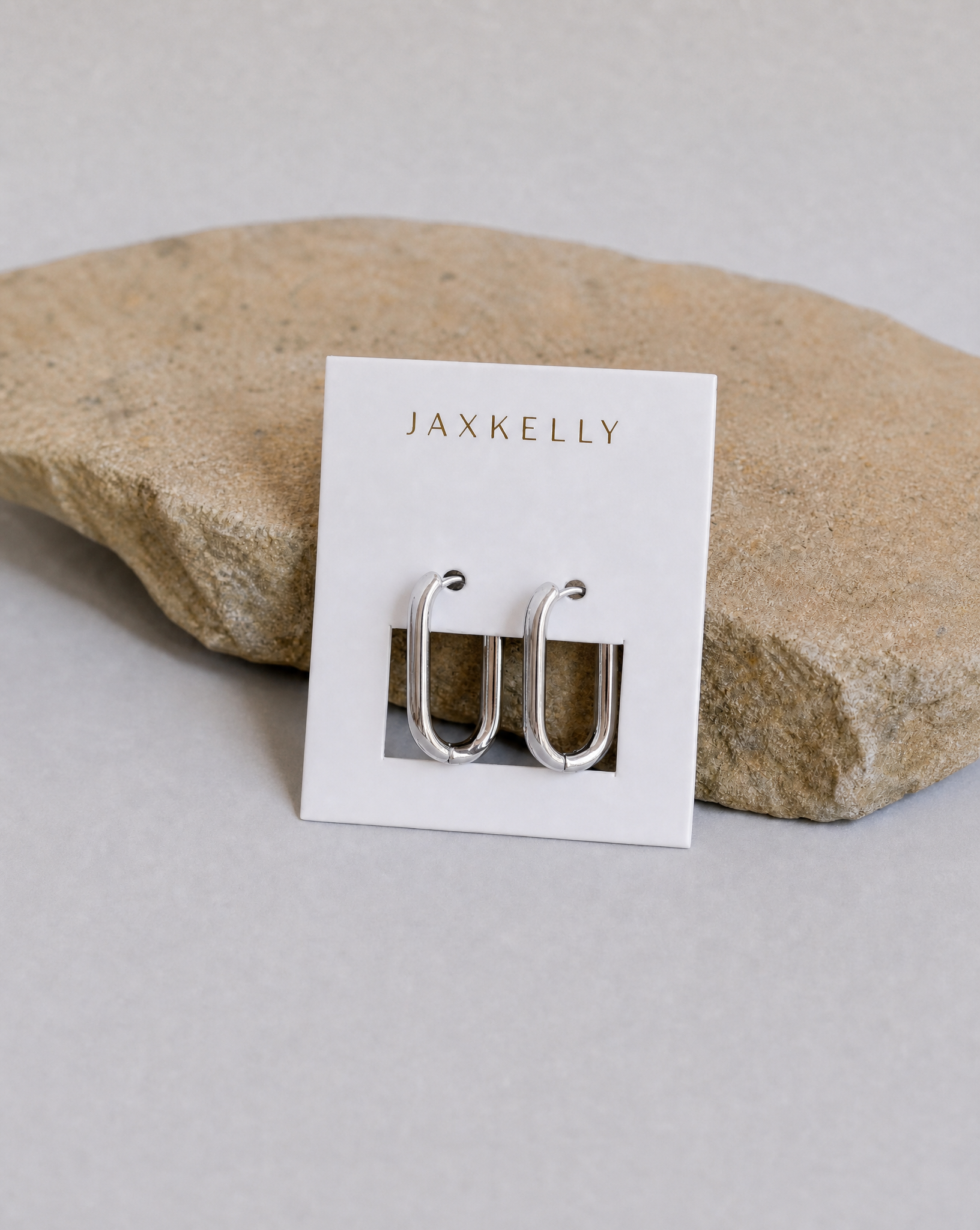 The JaxKelly Silver Rectangle Hoop - Large earrings, featuring sterling silver posts, are shown on a white branded card placed on a textured light brown stone with a neutral backdrop.