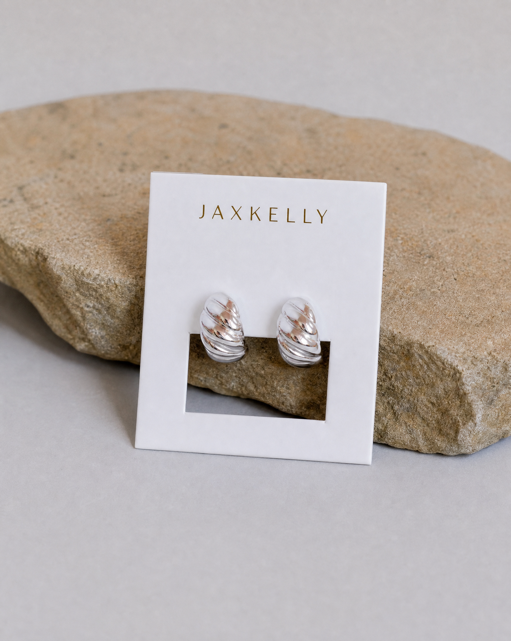 The JaxKelly Silver Swirl Hoop earrings, featuring a subtle twisted design, are shown on a branded white card against a textured beige stone background.