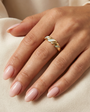 A close-up of a hand with light pink manicured nails wearing the Gold Twist Ring - White Opal, featuring a lab-grown opal inlay, resting on soft beige fabric.