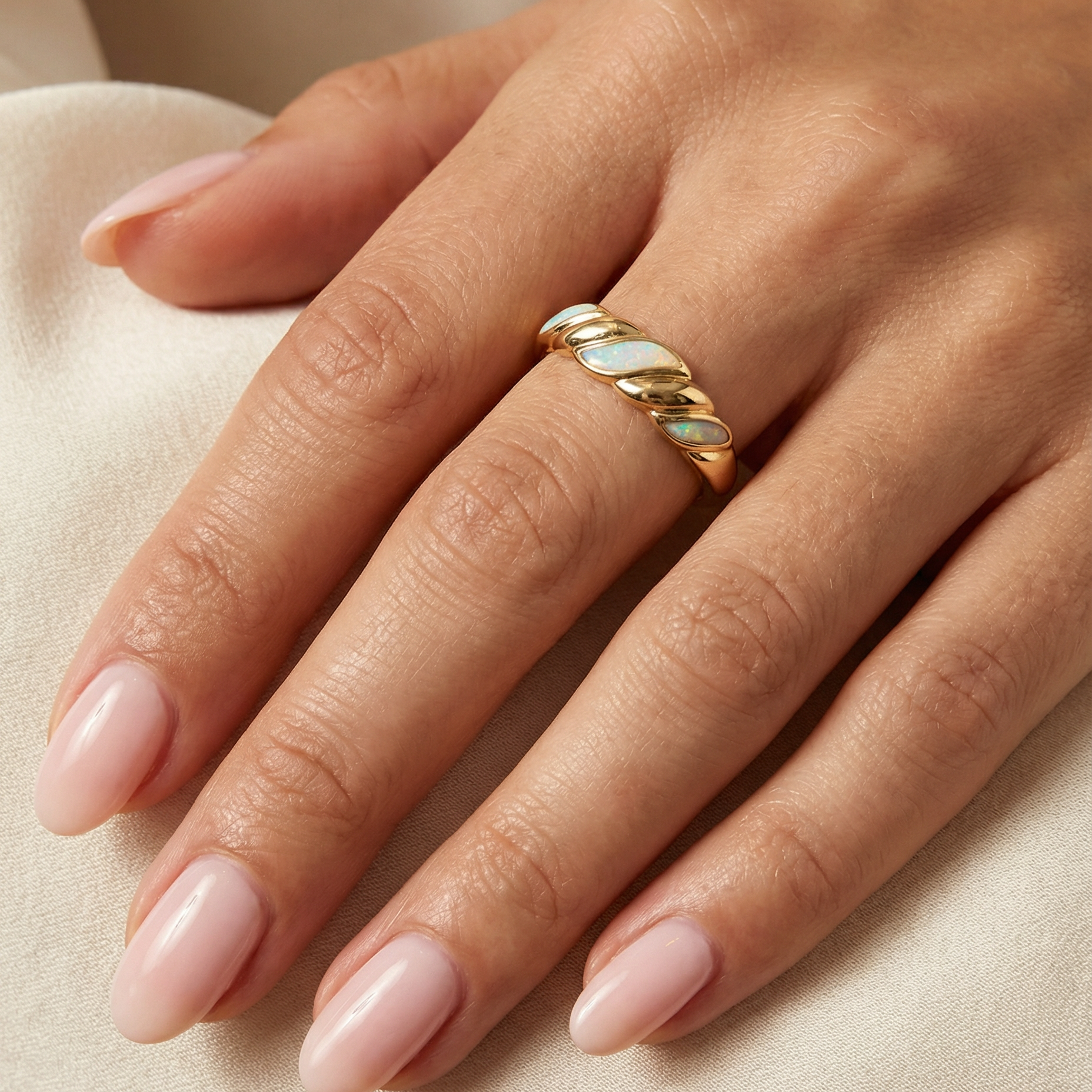 A close-up of a hand with light pink manicured nails wearing the Gold Twist Ring - White Opal, featuring a lab-grown opal inlay, resting on soft beige fabric.