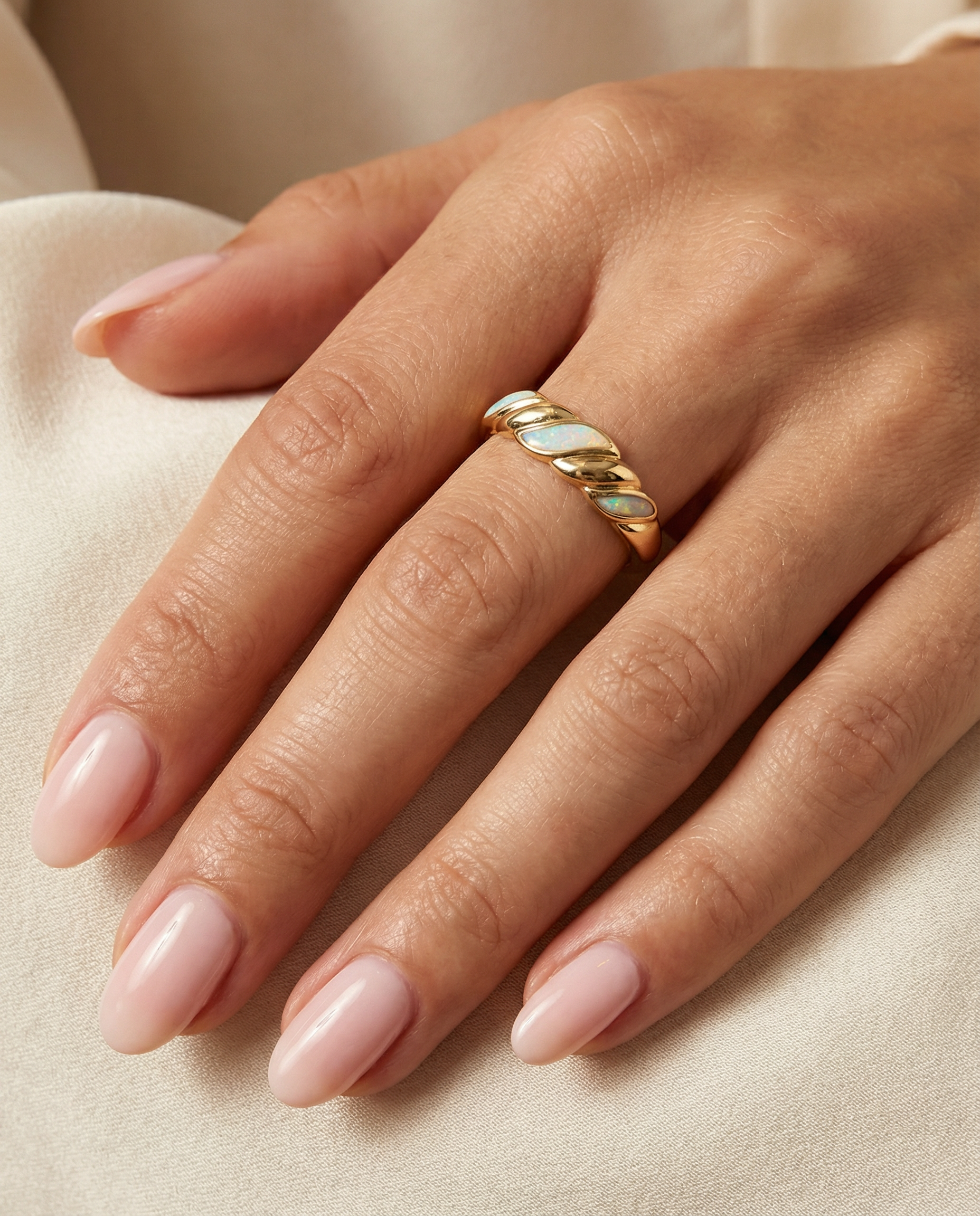 A close-up of a hand with light pink manicured nails wearing the Gold Twist Ring - White Opal, featuring a lab-grown opal inlay, resting on soft beige fabric.