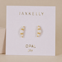 The Crescent Gold Earrings - White Opal by JaxKelly, featuring lab-grown opals and 18K gold plating, are displayed on a white card with “JAXKELLY” at the top and “OPAL joy” at the bottom, set against a neutral background.