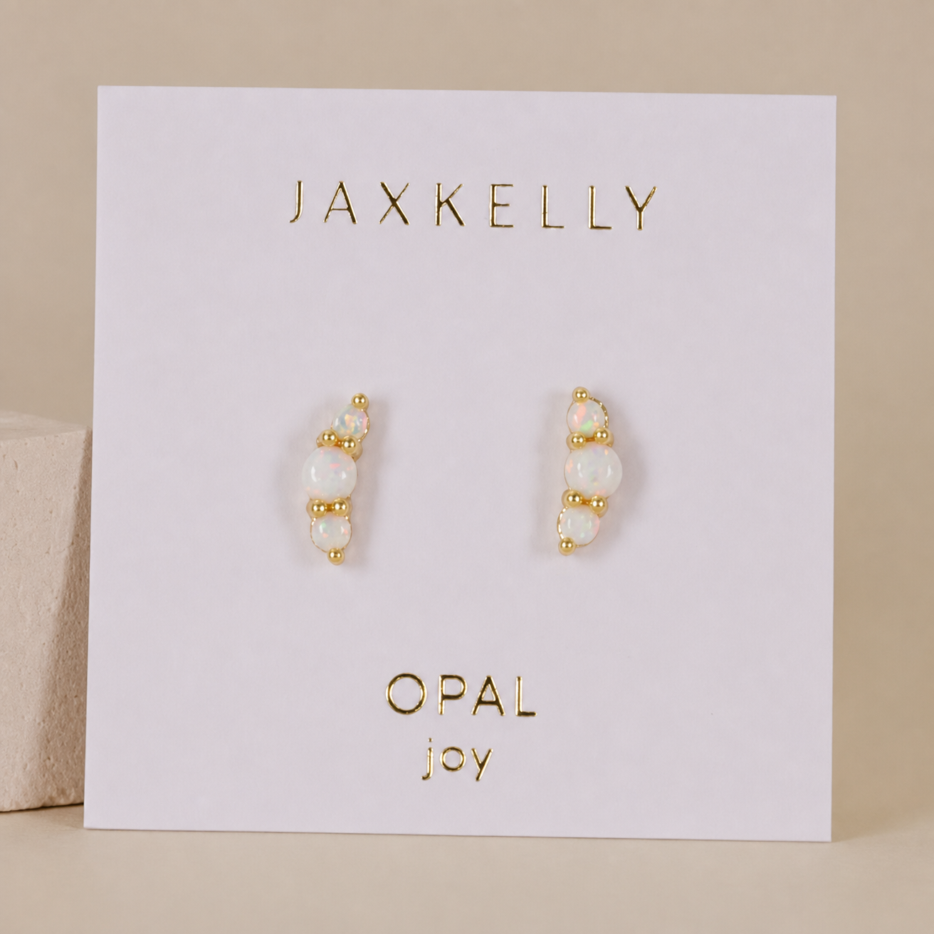 The Crescent Gold Earrings - White Opal by JaxKelly, featuring lab-grown opals and 18K gold plating, are displayed on a white card with “JAXKELLY” at the top and “OPAL joy” at the bottom, set against a neutral background.