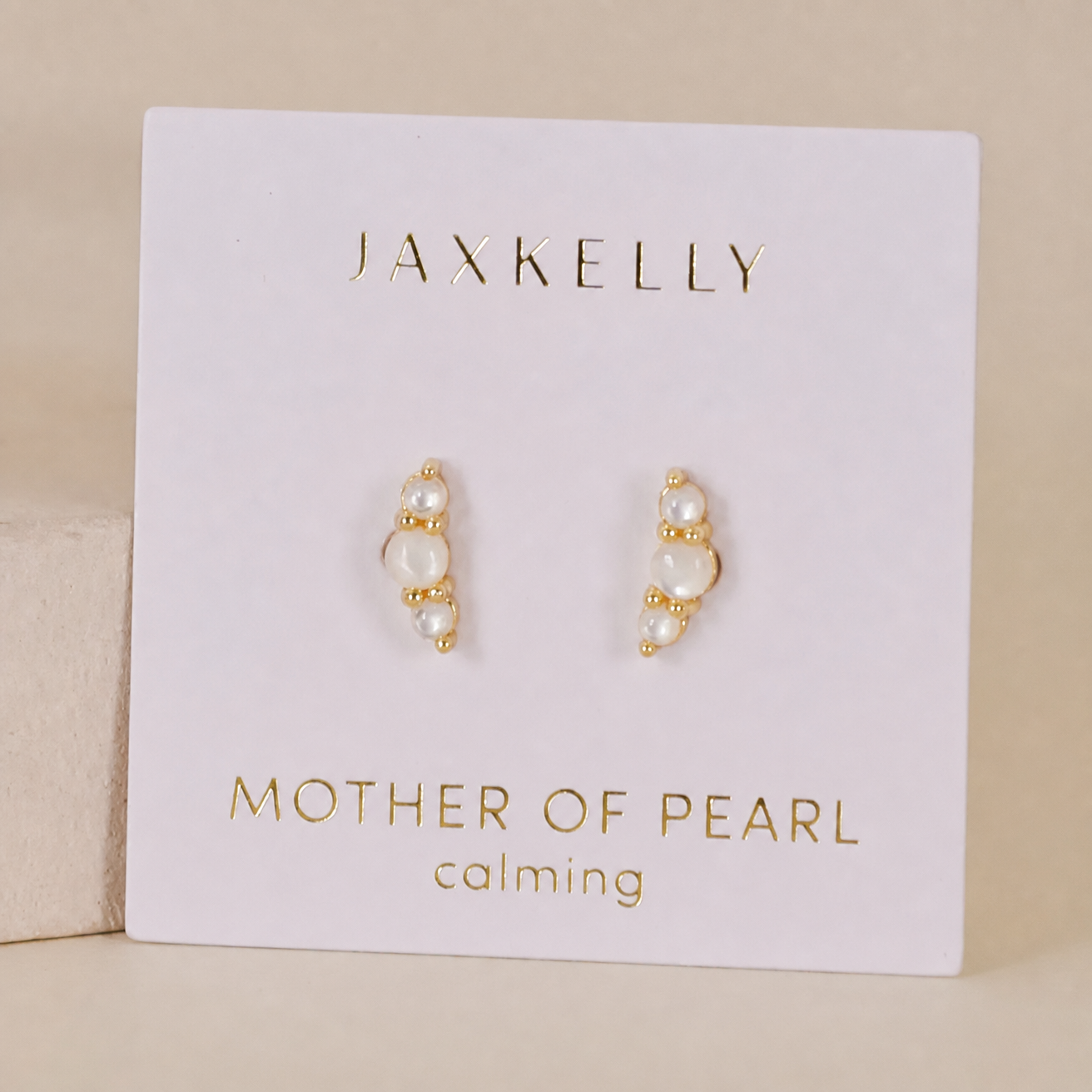 A pair of Crescent Gold Earrings - Pearl by JaxKelly on a white card reading "Mother of Pearl" and "calming" in gold. Each features small, vertically aligned pearls with comfortable sterling silver posts.