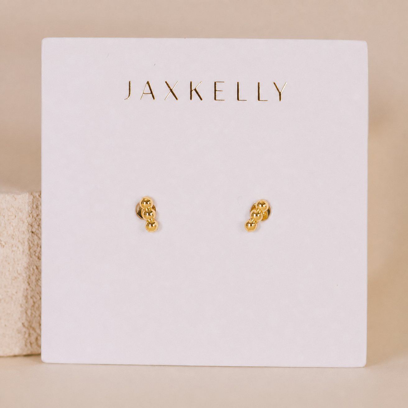 The JaxKelly Petite 3 Ball Stud earrings feature a small, gold cluster design with gold plating. Displayed on a white card with JaxKelly in gold lettering, these chic minimalist studs are also hypoallergenic—perfect for everyday wear.
