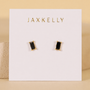 The JaxKelly Black Baguette earrings feature rectangular black studs with 18k gold plating and sterling silver posts, displayed on a white branded card against a beige background.