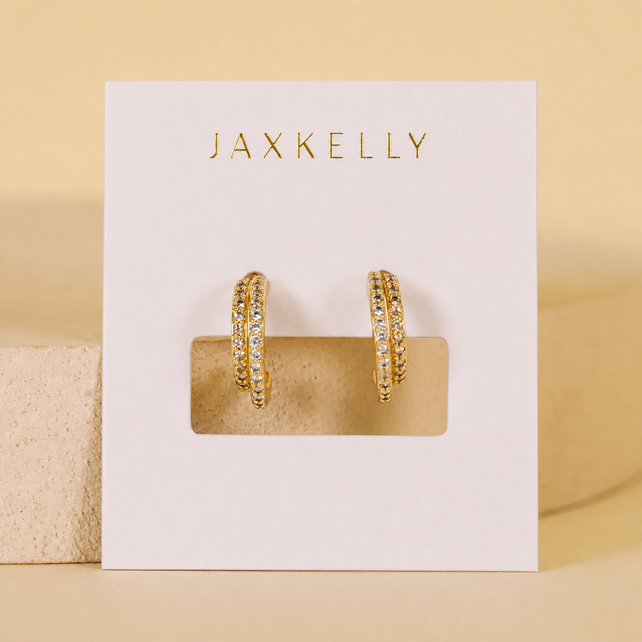 The JaxKelly Double Champagne Hoop Earrings feature double offset hoops with clear pavé stones and 18K gold plating, displayed on a white JaxKelly card against a beige background.
