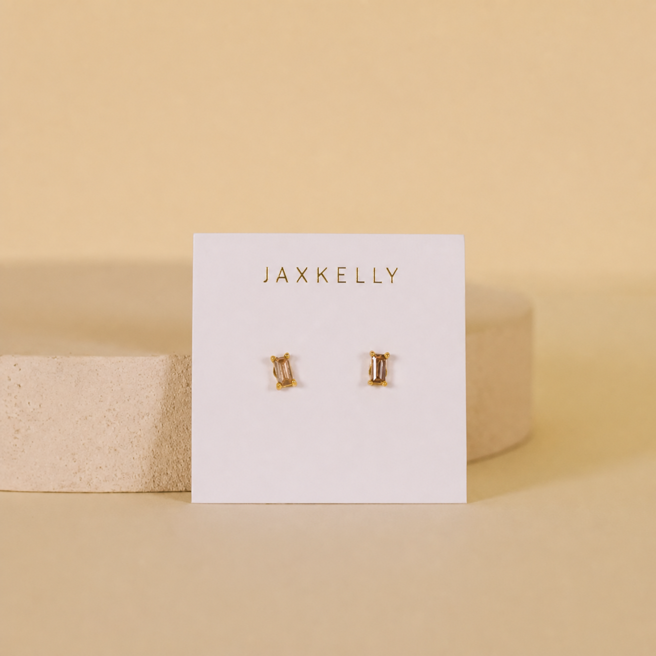 A pair of Coffee Baguette gold-plated rectangle stud earrings by JaxKelly, displayed on a white card with gold lettering, set against a beige background with a round stone prop. Hypoallergenic and perfect for daily wear.